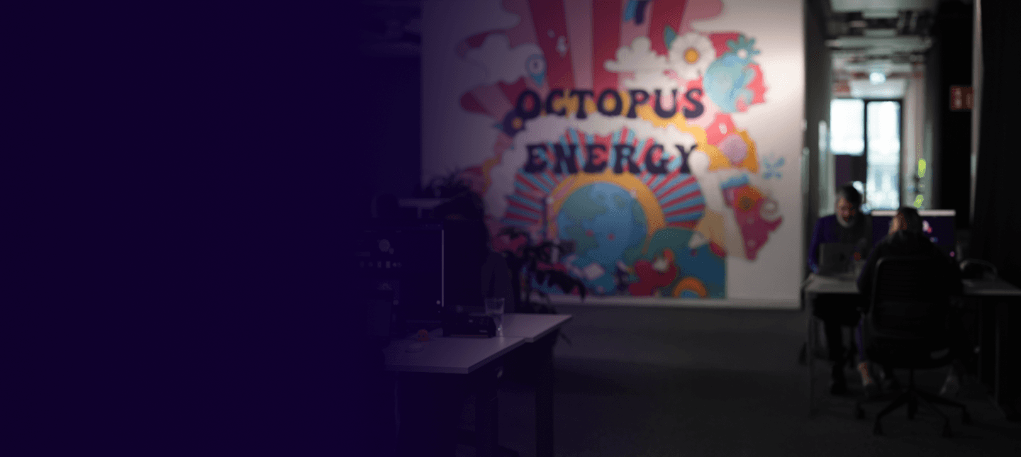 A dimly lit office space with a colorful Octopus Energy mural on the wall. Two people work at desks near a window.