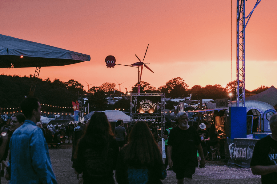 Menschen, die bei Sonnenuntergang auf einem Open-Air-Festival spazieren gehen, im Hintergrund Zelte, Banner und eine Windkraftanlage.