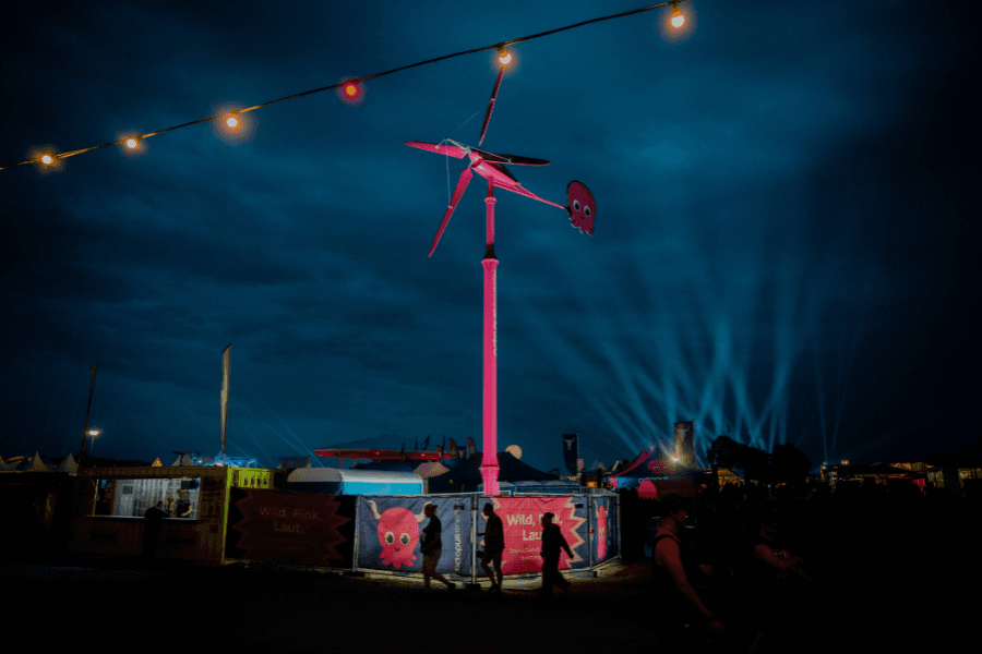 Eine rosa Windkraftanlage leuchtet nachts in einer festlichen Kulisse, im Vordergrund sind bunte Lichter und die Silhouetten von Menschen zu sehen.